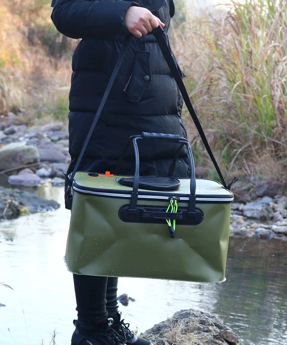 Una persona parada cerca de una masa de agua, posiblemente un río o arroyo, con una hielera verde en sus manos. La hielera tiene un asa negra y parece estar equipada con una correa para el hombro para transportarla.