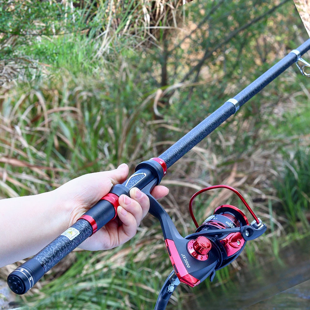 Una persona que sostiene una caña de pescar con un carrete adjunto. La caña parece ser una caña giratoria, que se usa comúnmente para varios tipos de pesca, incluida la de agua dulce y salada. El carrete tiene una combinación de colores rojo y negro, y la caña tiene un mango negro con un detalle en rojo.