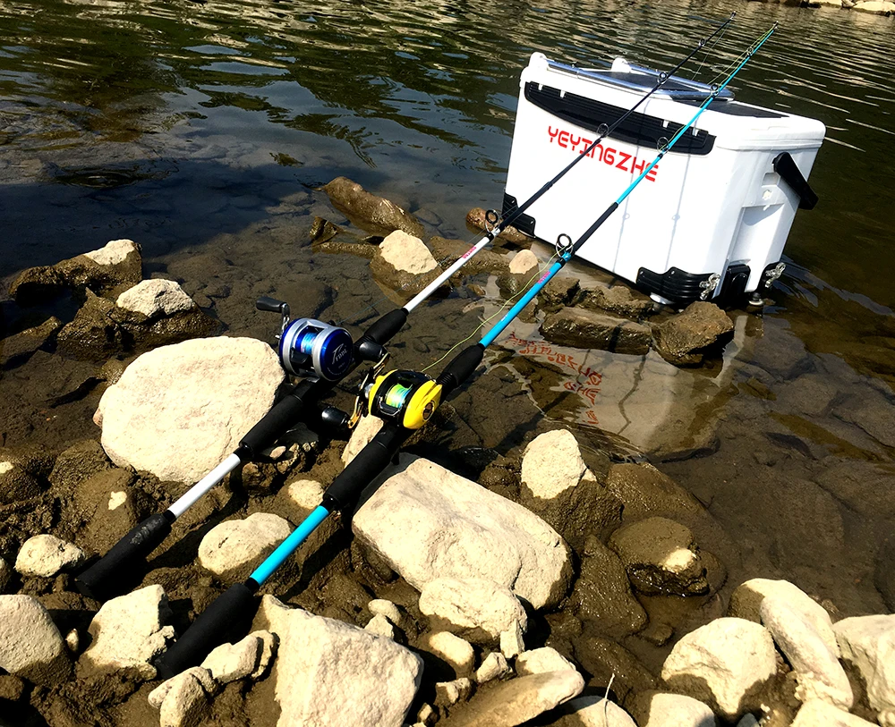 Un establecimiento de pesca en una costa rocosa. Hay una hielera blanca con la marca YETI visible, que es una marca popular conocida por sus hieleras aisladas. Al lado de la hielera, hay dos cañas de pescar con carretes adjuntos, apoyadas sobre las rocas.