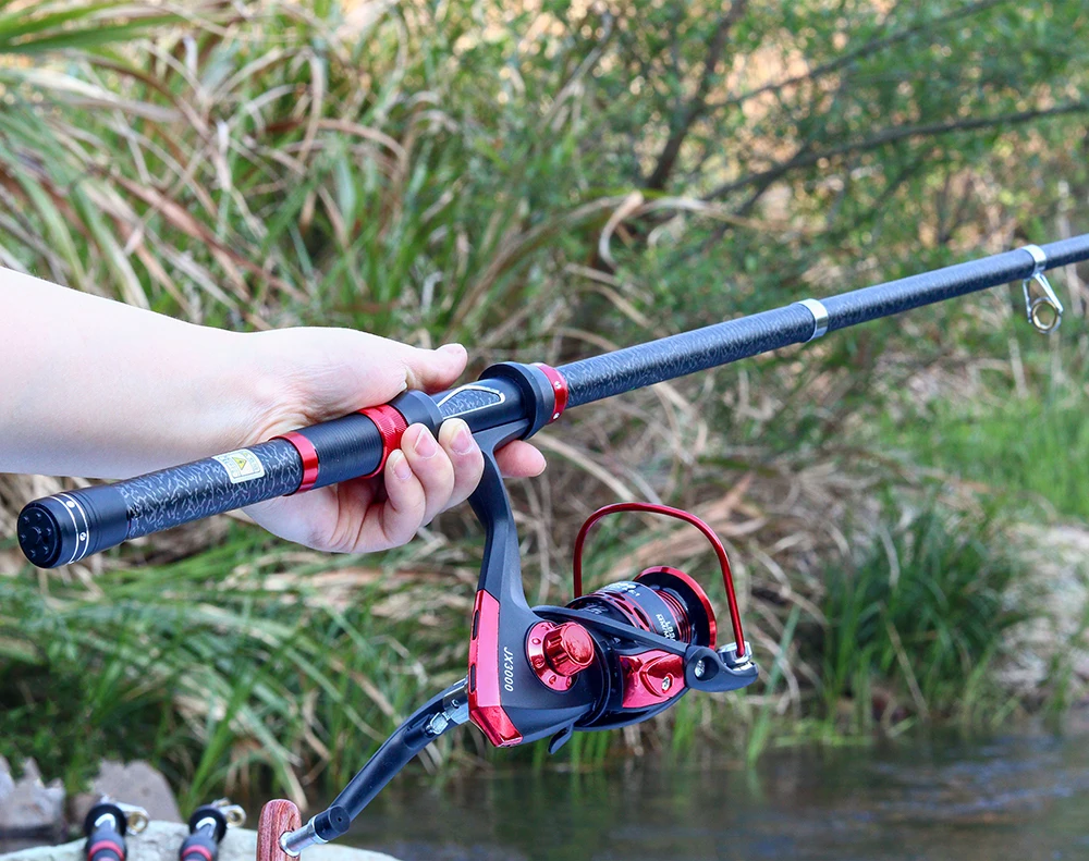 Una persona que sostiene una caña de pescar con un carrete adjunto. La caña parece ser una caña giratoria, que se usa comúnmente para la pesca en agua dulce. El carrete tiene un esquema de color rojo y negro, y la caña también tiene un diseño negro y rojo.