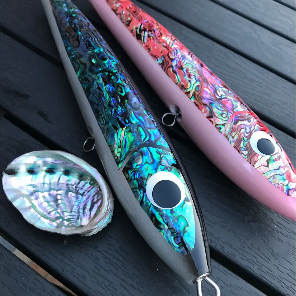 Dos señuelos de pesca con una concha de perla en primer plano. Los señuelos tienen un cuerpo metálico con un patrón colorido e iridiscente que se asemeja a las escamas de un pez. Tienen un anzuelo al final y un pequeño ojal para sujetarlo a un hilo de pescar.