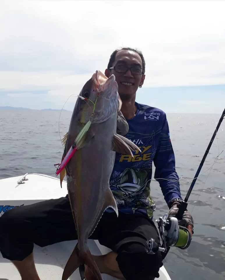 Una persona sentada en un barco, sosteniendo un pez grande. El pez parece ser un tipo de pez picudo, posiblemente un pez vela o un marlín, dado su tamaño y la presencia de un pico. La persona lleva una camisa azul con un gráfico y gafas.