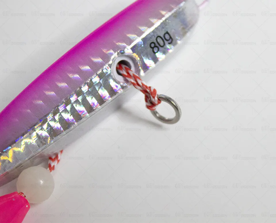 Un primer plano de un objeto rosa con brillo metálico, posiblemente una joya o un elemento decorativo. Tiene una pequeña cuenta redonda y blanca adherida y una cuerda o cinta con rayas rojas y blancas está atada alrededor de la cuenta.