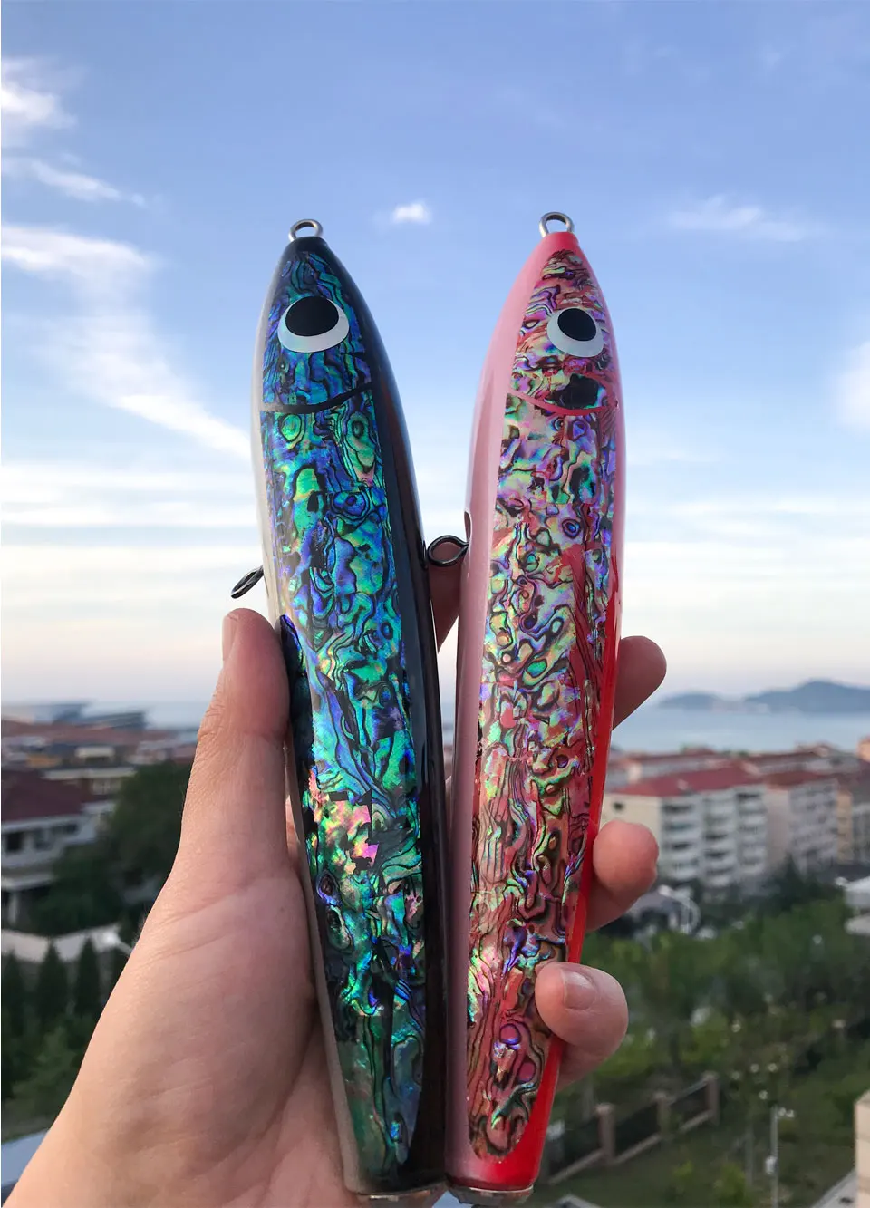 Dos manos sosteniendo lo que parecen ser lápices o bolígrafos. Los lápices tienen un acabado metálico con un patrón que se asemeja a una escama de pez o un diseño iridiscente similar. El de la izquierda tiene un tono azulado, mientras que el de la derecha tiene un tono rosado.