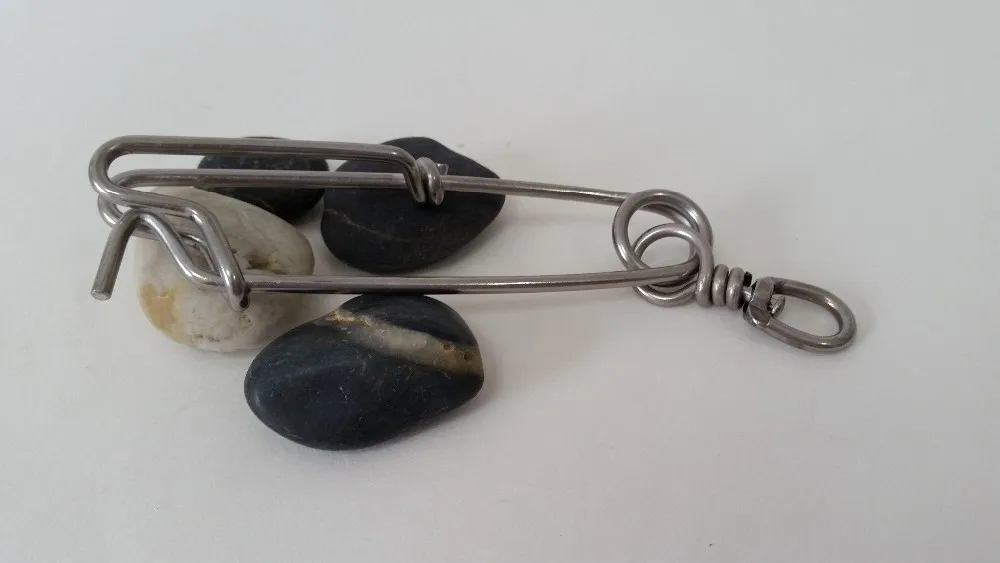 Un mosquetón de metal con un llavero adjunto, colocado sobre una superficie con tres rocas. El mosquetón parece estar hecho de un metal plateado, posiblemente acero o aluminio, y tiene una puerta con resorte que se puede abrir para colocar o quitar artículos.