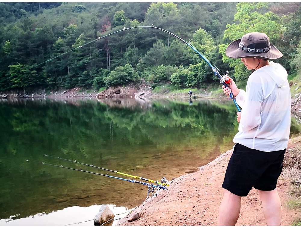 Una persona que pesca en una masa de agua tranquila, probablemente un lago o un río de corriente lenta. La persona lleva un sombrero y una camisa de color claro, y sostiene una caña de pescar con un hilo extendido hacia el agua.