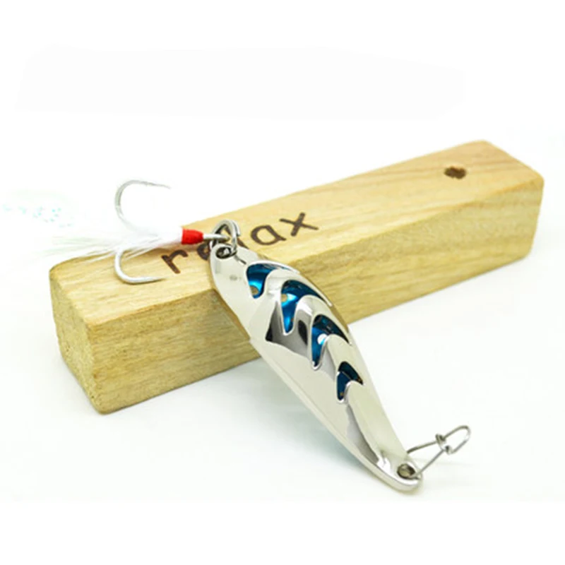 Un señuelo de pesca con un anzuelo adherido, apoyado sobre un bloque de madera. El señuelo tiene un cuerpo metálico con una combinación de colores azul y plateado, y parece estar diseñado para parecerse a un pez o un pez cebo.