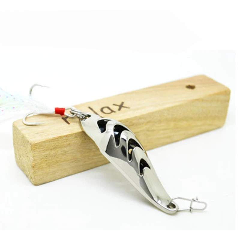 Un señuelo de pesca con un anzuelo rojo, colocado sobre un bloque de madera. El señuelo tiene un acabado metálico brillante y parece estar diseñado para parecerse a un pez o un pez carnada, lo cual es típico de los señuelos utilizados en la pesca para atraer peces.