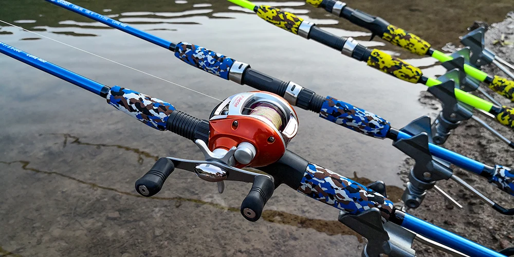 Un carrete de pesca unido a una caña de pescar. El carrete es naranja y plateado, y la caña tiene un patrón de camuflaje con colores azul y negro. El hilo de pescar es visible y se extiende desde el carrete hasta el agua.