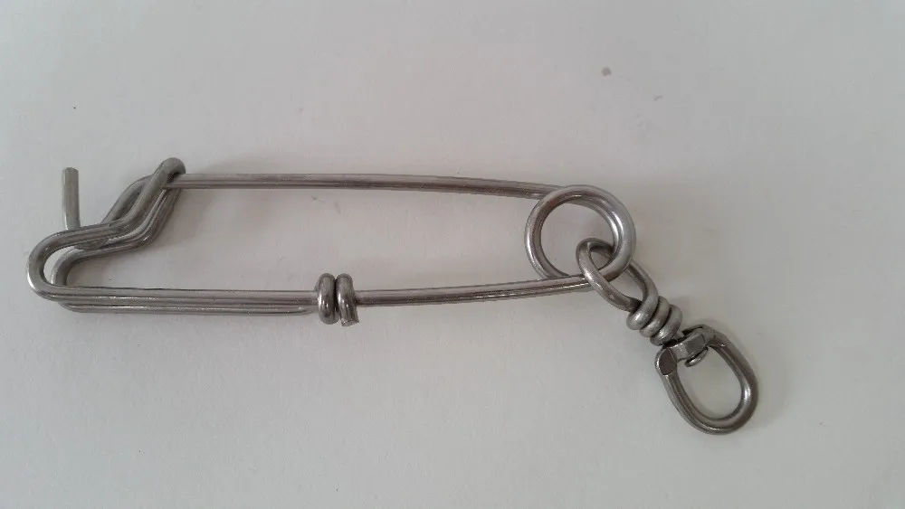 Un objeto metálico que parece ser una especie de gancho o clip. Tiene un lazo en un extremo y un gancho en el otro, con una cadena o eslabón que conecta los dos. Este tipo de objeto se utiliza a menudo para asegurar elementos juntos o para colgar cosas.