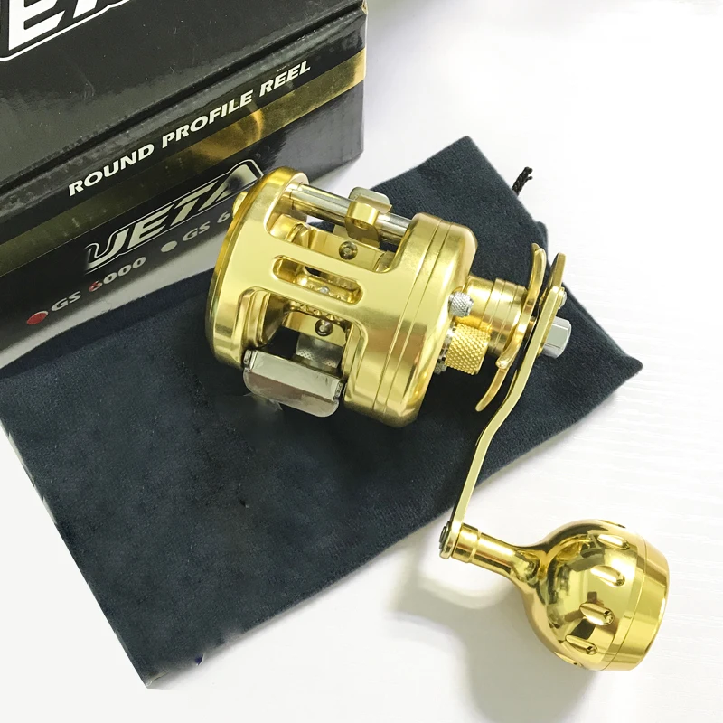 Un carrete de perfil redondo de color dorado, que es un tipo de carrete de pesca. Parece ser un carrete nuevo o sin usar, como lo indica el empaque y el hecho de que está colocado sobre un paño, posiblemente con fines de protección o exhibición.