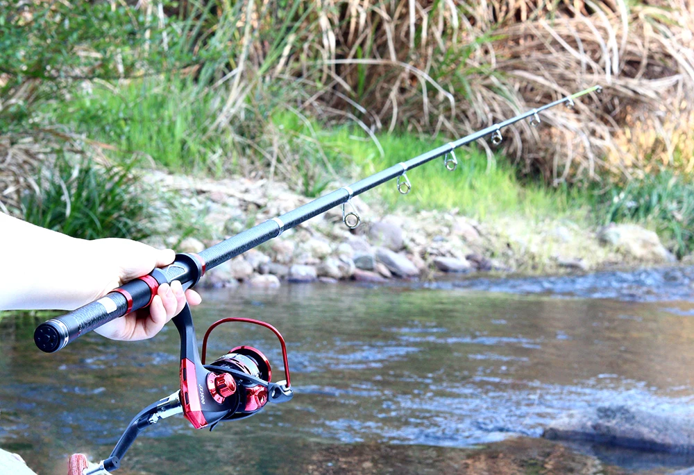 Una persona que sostiene una caña de pescar con un carrete adjunto. La caña se extiende y la persona parece estar lanzando o recuperando una línea. El fondo sugiere que la persona está pescando en un entorno natural, posiblemente cerca de un río o arroyo, con vegetación y rocas visibles.