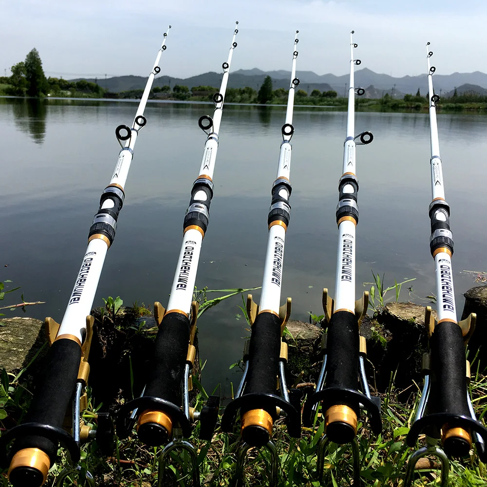 Una colección de cañas de pescar apoyadas contra una roca o una estructura cerca de una masa de agua, que parece ser un lago o un río tranquilo. Las cañas están dispuestas en fila, con sus mangos apuntando hacia el espectador y sus puntas extendiéndose hacia el agua.