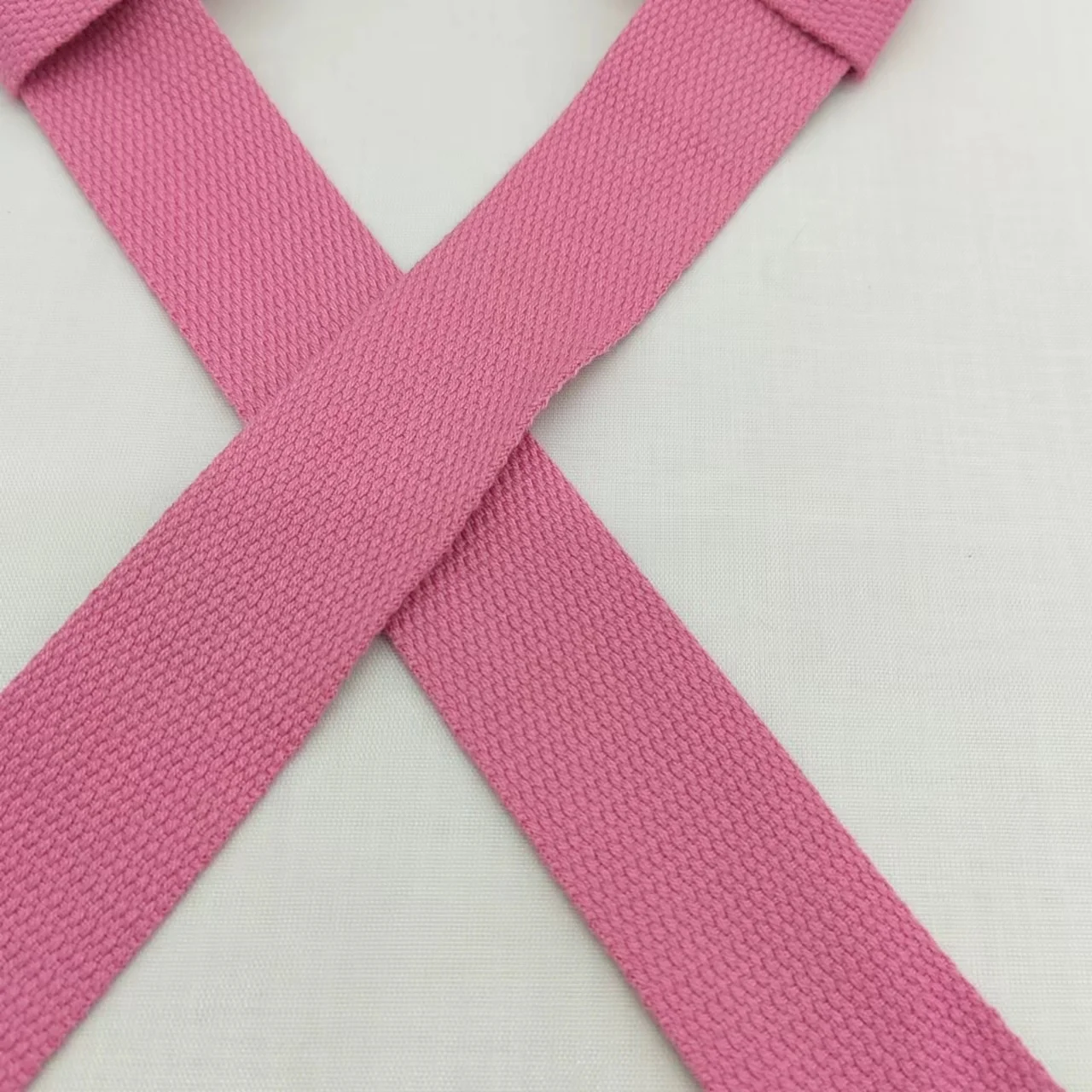 Una cinta rosa atada en forma de X sobre un fondo blanco. La cinta parece estar hecha de una tela tejida y se asocia comúnmente con la concienciación sobre el cáncer de mama, ya que las cintas rosas se utilizan a menudo como símbolo de apoyo a las personas afectadas por la enfermedad.