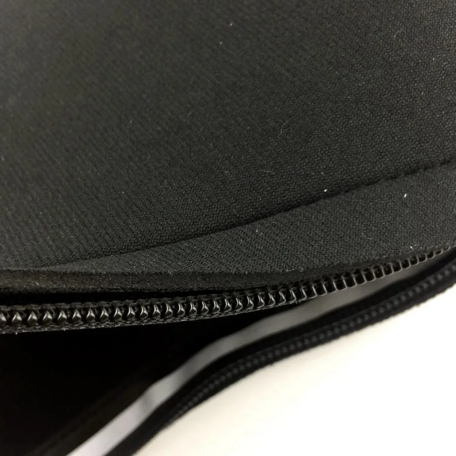 Un primer plano de una tela negra con detalle de cremallera. Se ve parcialmente la cremallera, con sus dientes y el borde de la tela. La tela parece ser de un color sólido y es probable que la cremallera se use para abrir o cerrar una prenda o un bolso.