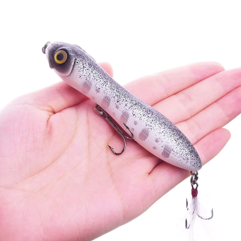 La mano de una persona sosteniendo un señuelo de pesca. El señuelo está diseñado para parecerse a un pez, con un cuerpo con un patrón moteado, un ojo amarillo y un anzuelo sujeto al extremo de la cola.