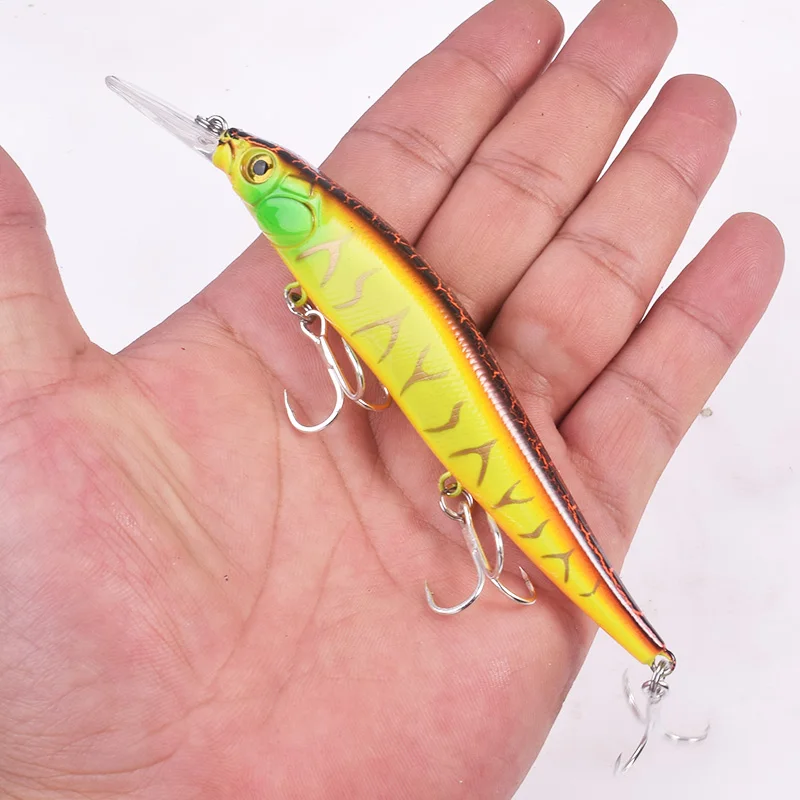 Una mano sosteniendo un señuelo de pesca. El señuelo está diseñado para parecerse a un pez y normalmente se utiliza para atraer peces reales cuando se pesca. Tiene un cuerpo con un patrón que imita las escamas de un pez y está equipado con anzuelos y un anillo partido para sujetarlo a un hilo de pescar.