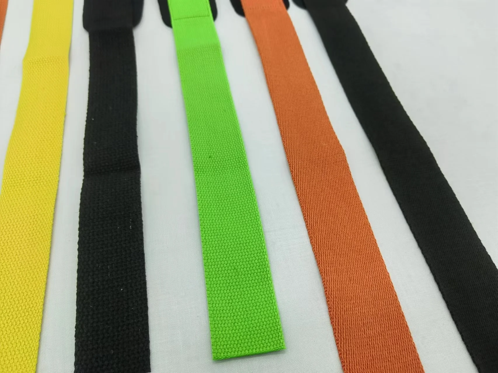 Una colección de cinturones de tela con diferentes colores. Hay cinturones negros, amarillos, verdes y naranjas, cada uno con una textura tejida. Las correas parecen estar dispuestas planas, posiblemente con fines de visualización o comparación.