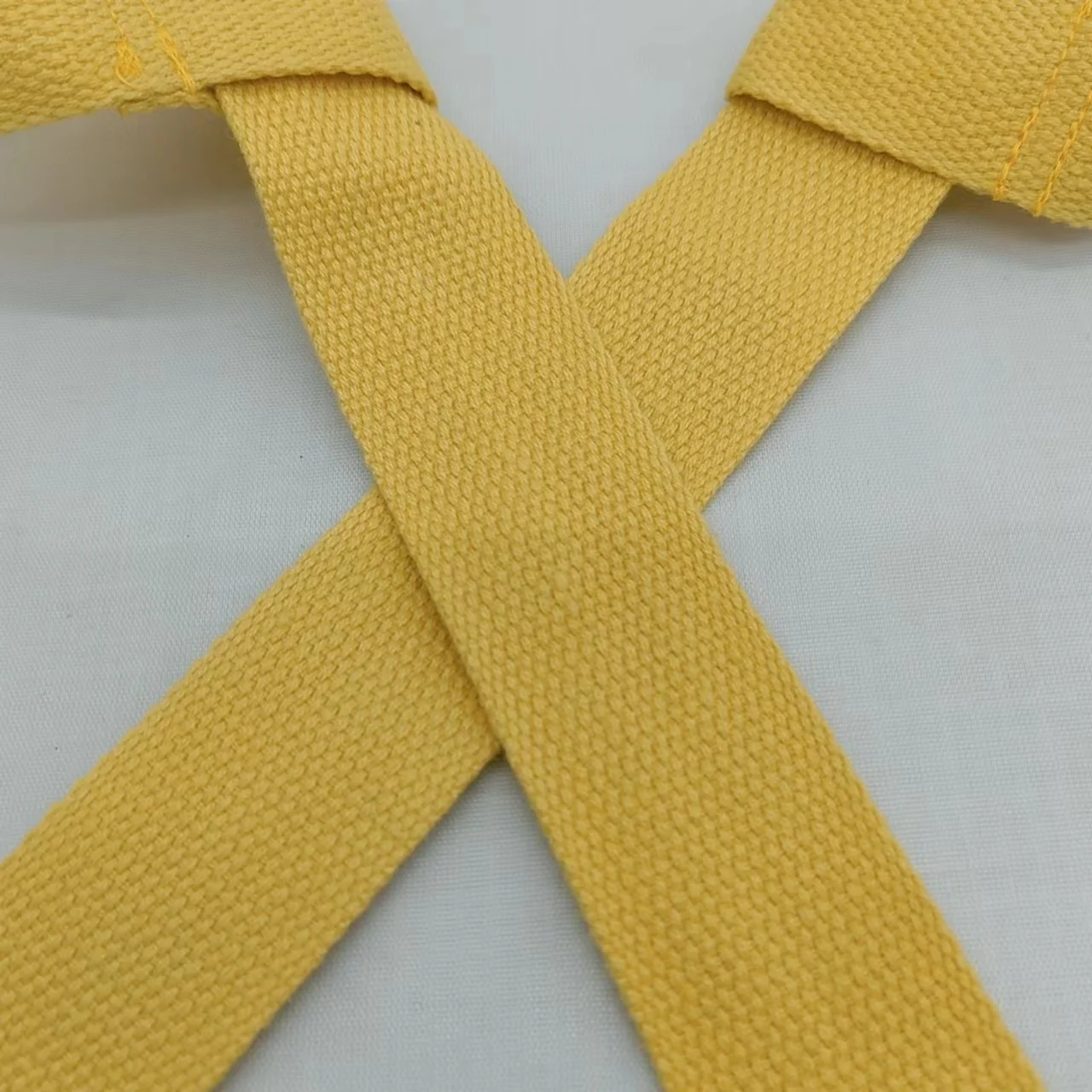 Un cinturón amarillo con textura tejida, extendido sobre una superficie blanca. El cinturón parece ser un tipo de cinturón de artes marciales, posiblemente para karate u otro arte marcial similar, dado el diseño y el color.