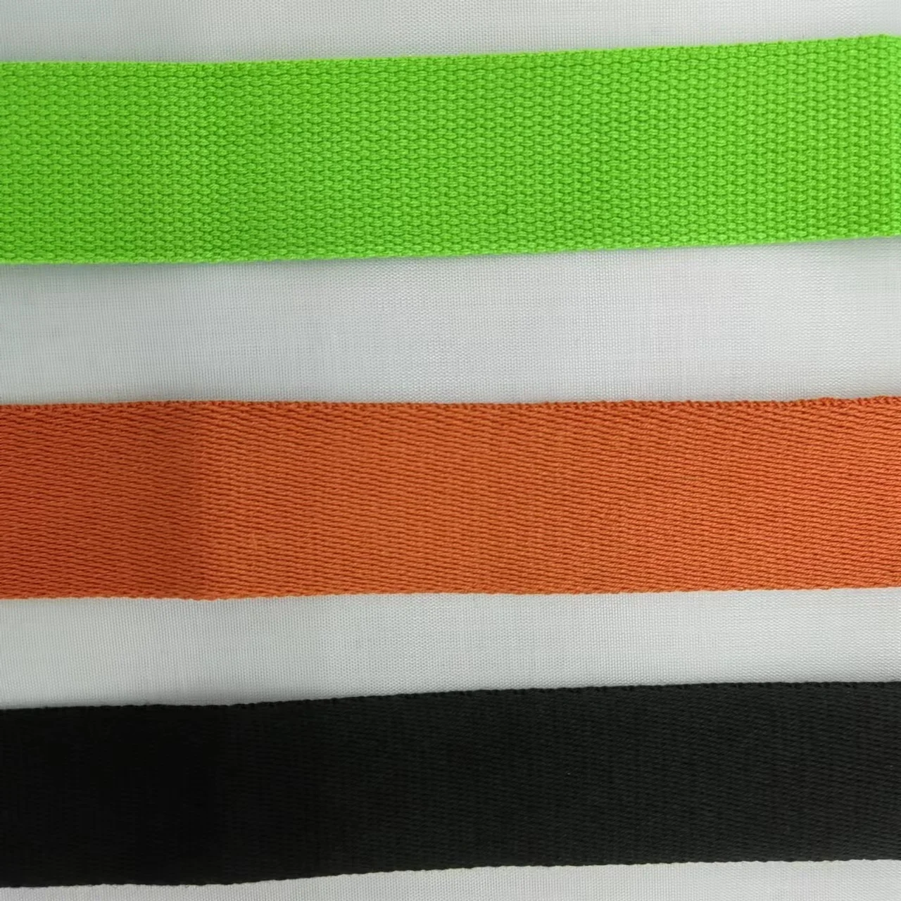 Tres bandas o tiras de tela sobre fondo blanco. De arriba a abajo, hay una banda verde, una banda naranja y una banda negra. Cada banda parece estar hecha de una tela tejida, posiblemente destinada a usarse como correa o corbata.