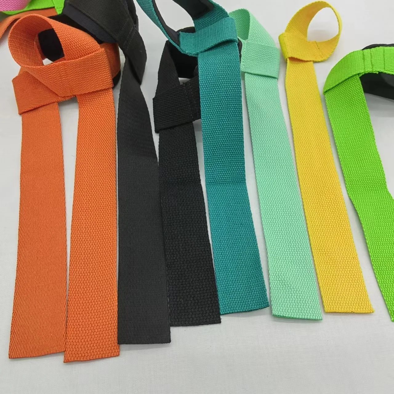 Colección de corbatas de tela en varios colores. Parecen estar hechos de un material tejido, posiblemente poliéster o un tejido sintético similar, y se muestran en fila con los extremos atados en un lazo. Los colores incluyen naranja, negro, verde azulado, verde y amarillo.