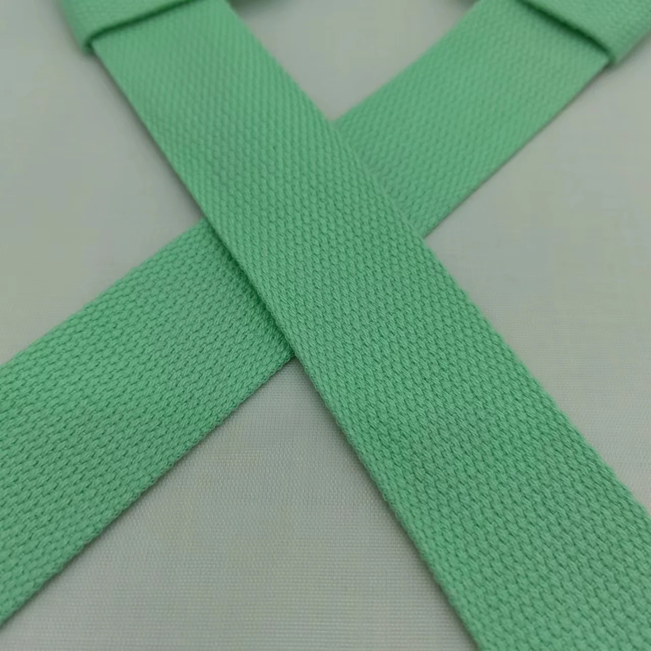 Un cinturón de tela verde con textura tejida. Parece ser un cinturón simple y plano con un extremo en forma de bucle, que generalmente se usa para sujetar prendas de vestir como pantalones o faldas.