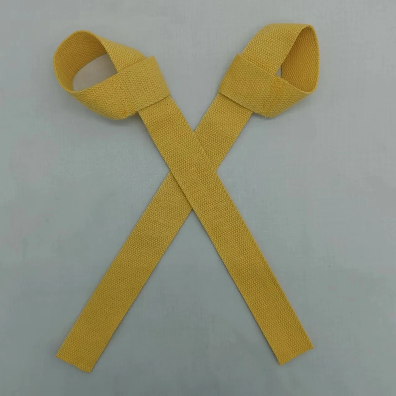Un cinturón de tela amarilla con textura tejida. Parece ser un cinturón simple y plano con un lazo en un extremo, que probablemente se usa para asegurar el cinturón alrededor de la cintura u otra parte del cuerpo. El cinturón se presenta plano sobre un fondo de color claro.