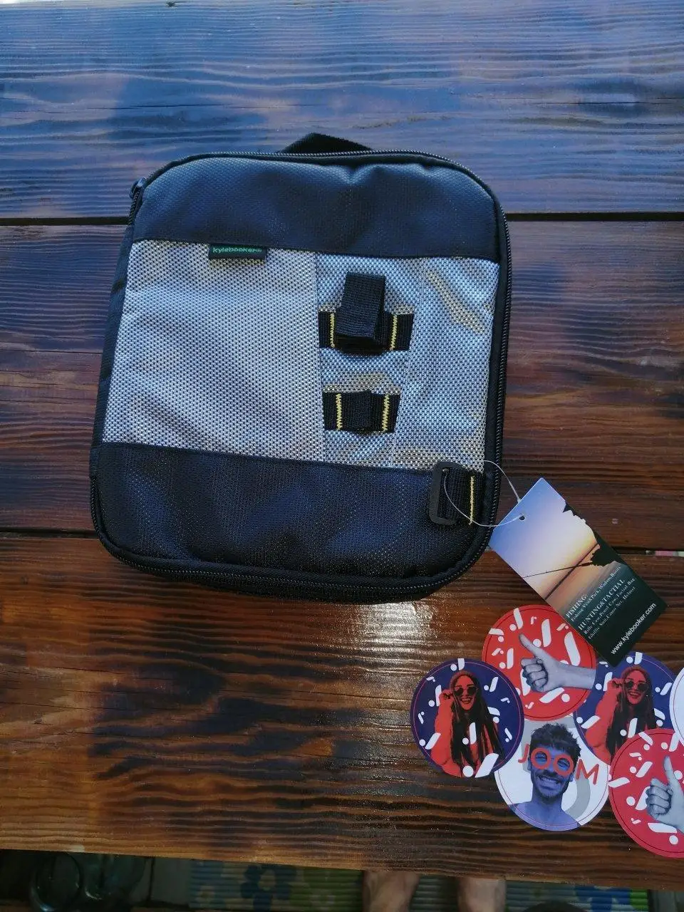 Un bolso tipo mensajero negro y gris con un compartimento con cremallera en la parte delantera. También hay varios elementos circulares sobre la mesa, que parecen ser pegatinas o insignias.