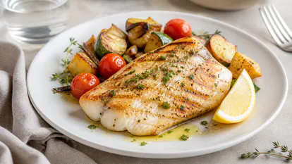 Plato de pescado jugoso presentado con verduras, limón y hierbas frescas.