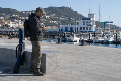 Pescador consulta los permisos en el móvil con la caña guardada en un paseo portuario