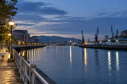 Recreación de una ría urbana inspirada en Bilbao con muelles iluminados al anochecer