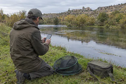 Pescador consultando la normativa en el móvil antes de utilizar sus reteles junto a un río