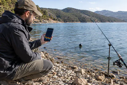 Pescador consultando el móvil junto a un sonar portátil flotante en la orilla de un embalse